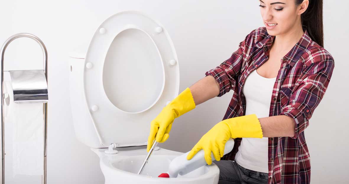 An image of a woman in a plaid shirt and yellow rubber gloves pouring bleach into a toilet bowl whilst cleaning it with a toilet brush. Her nose is crinkled from the smell.