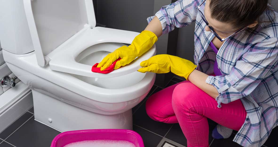 An image of a woman kneeling next to a white toilet. She is wearing yellow rubber gloves and is cleaning the toilet seat with a pink cloth.