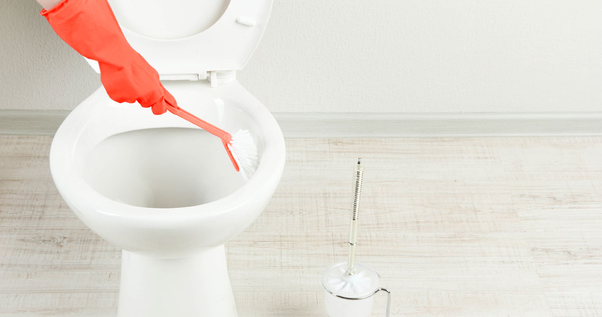 An image of an arm wearing a red rubber glove, cleaning the inside of a toilet bowl with a red scrub brush. There is a toilet brush to the left.