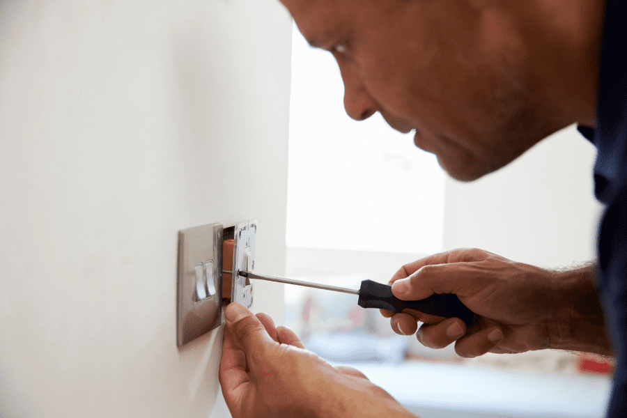 A close-up of an electrician's hand holding a screwdriver, undoing the screws on a light switch to replace it.